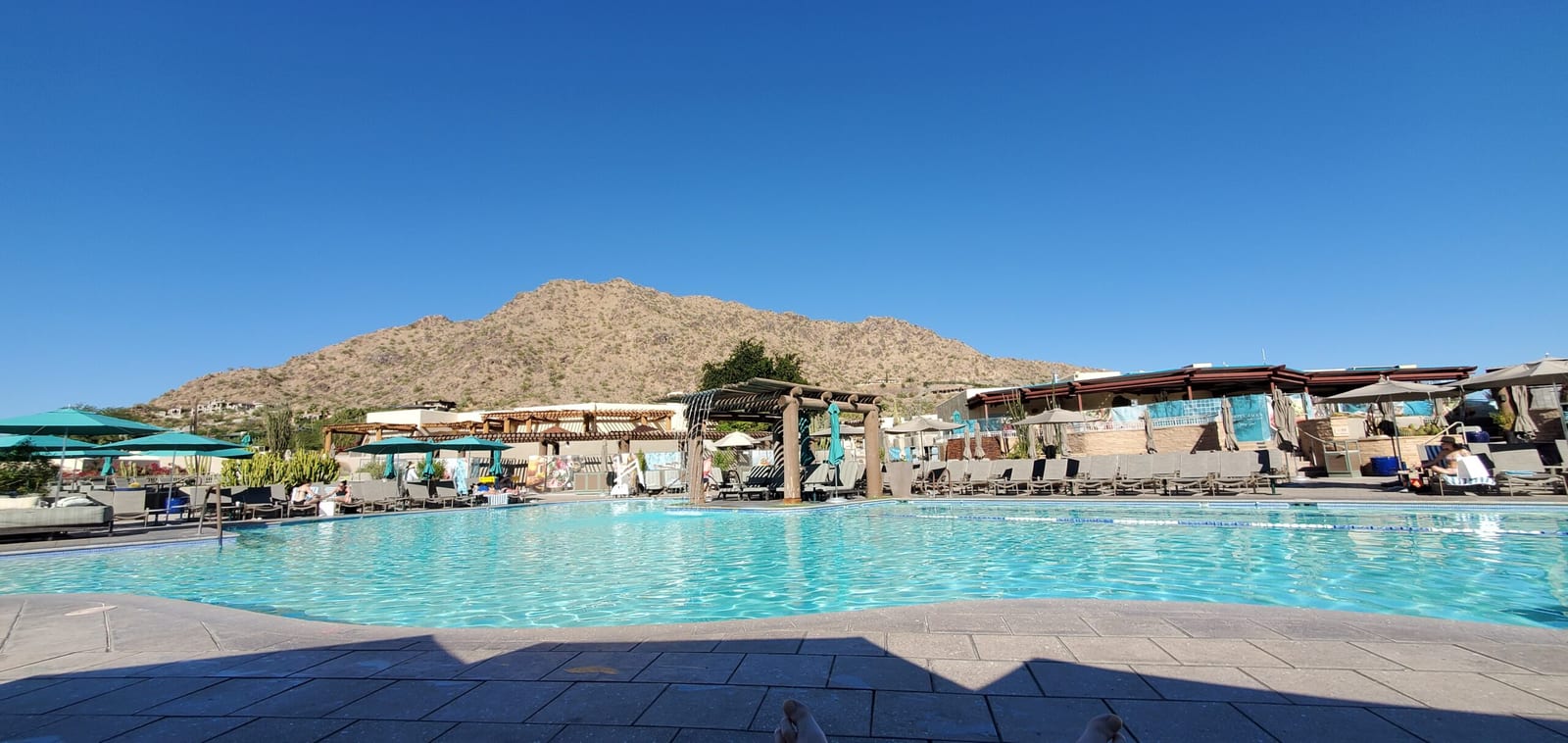 Sunbathers enjoying the heated Jackrabbit Pool with Mummy Mountain in the background. Daytime panorama of the Jackrabbit Pool under clear blue Arizona skies