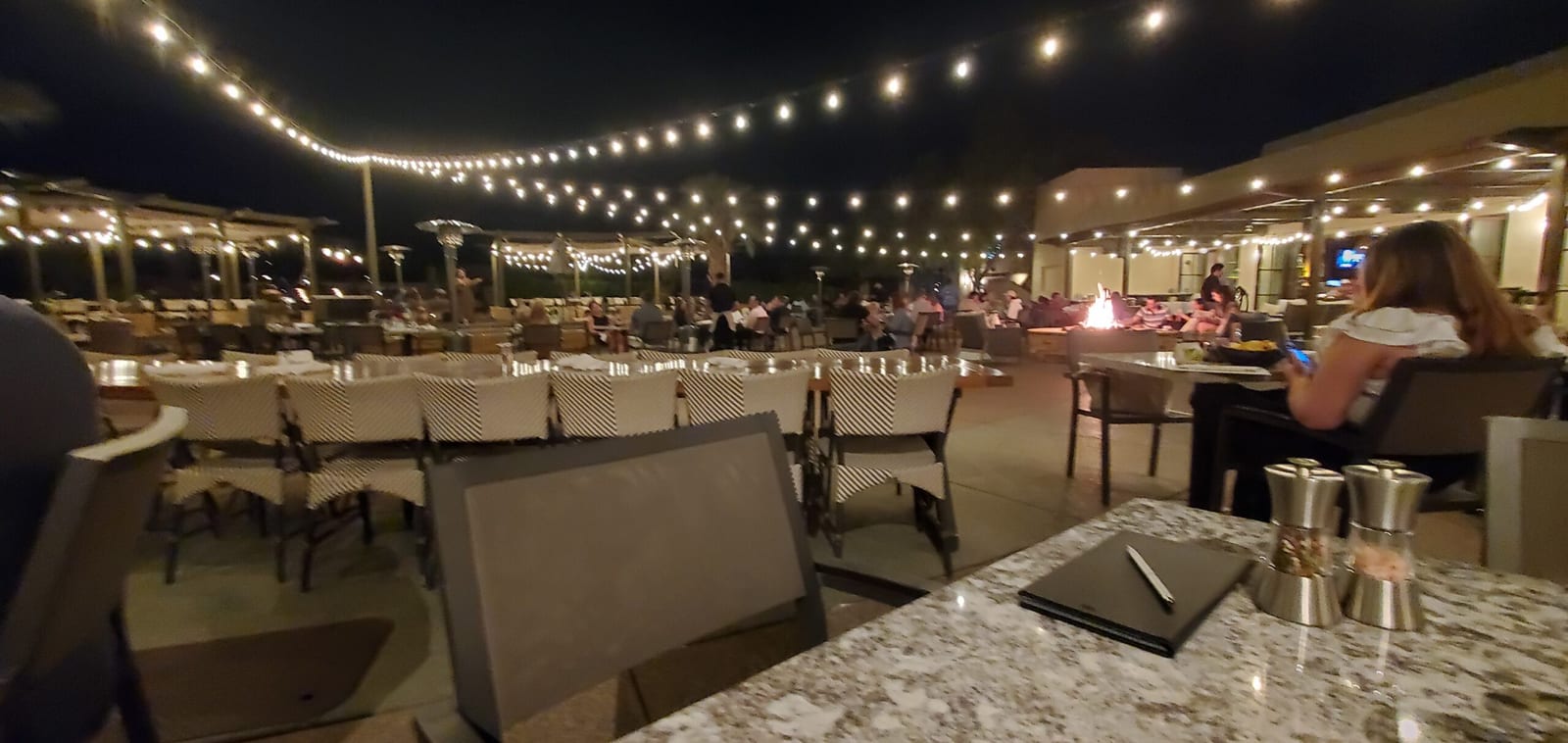 Evening dinner service on Rita's patio beneath bistro string lights and outdoor heaters. Rita's Cantina terrace dining under string lights with crackling fire pit